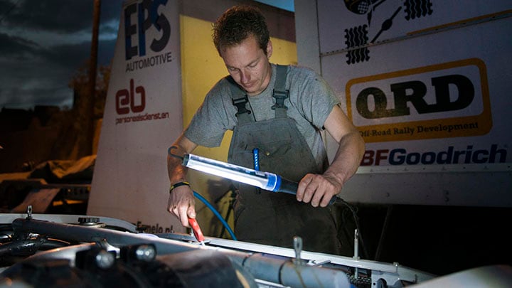 Un hombre se ayuda de una lámpara de trabajo LED para ver más claramente mientras repara un coche